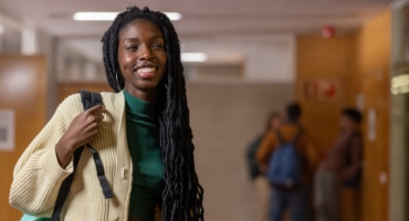 a woman smiling with a backpack on one shoulder standing in the hallway of a school