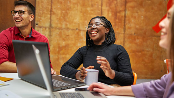 A group of young adults are sitting around a table with laptops, and they look like they are laughing and having a good time at work.