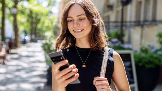A young woman is walking down a sunny street and looking at her phone with a smile on her face.