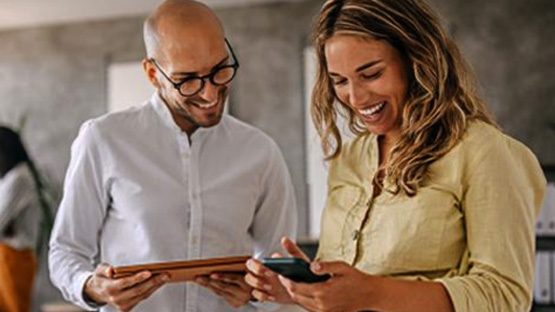 A bald man with glasses is smiling while looking at a phone that a woman with long blonde hair is holding, and they seem to be enjoying what they are seeing about their medical copays
