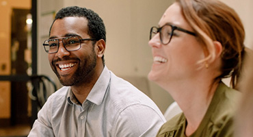 A group of people, including a man holding a white cup and a woman in glasses, are smiling in a meeting.