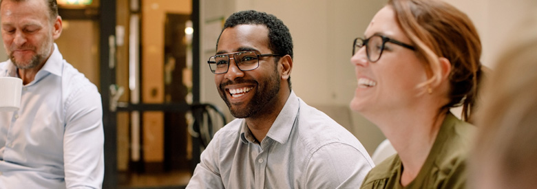 A group of people, including a man holding a white cup and a woman in glasses, are smiling in a meeting.