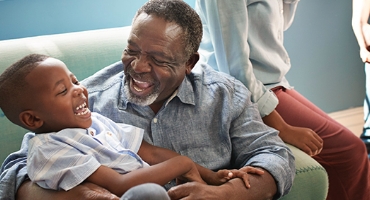 Black child sits on bearded grandfather's lap, both are laughing, someone nearby; represents Highmark Medicare Plans