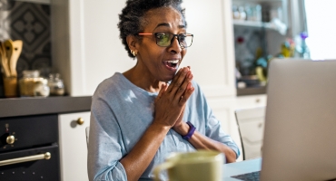  Older black woman sits at kitchen table at laptop, hands clasped, smiling and happily reading about Highmark Medicare Plans 