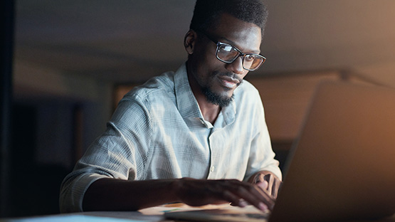 Black man with glasses browsing mental health resources on a laptop in a dark room.