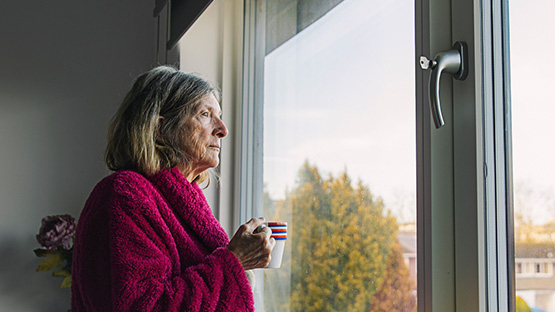 Older woman in bathrobe with a cup of coffee looking out the glass door