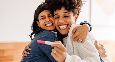two women hugging and smiling while holding a postive pregnancy test